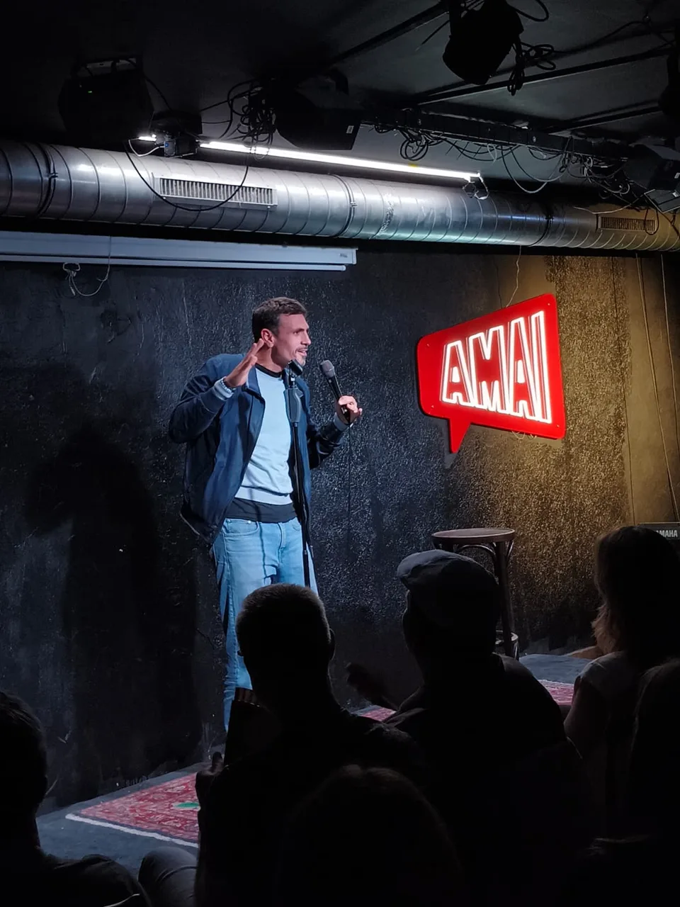 A comedian holding a microphone beside the illuminated Amai sign